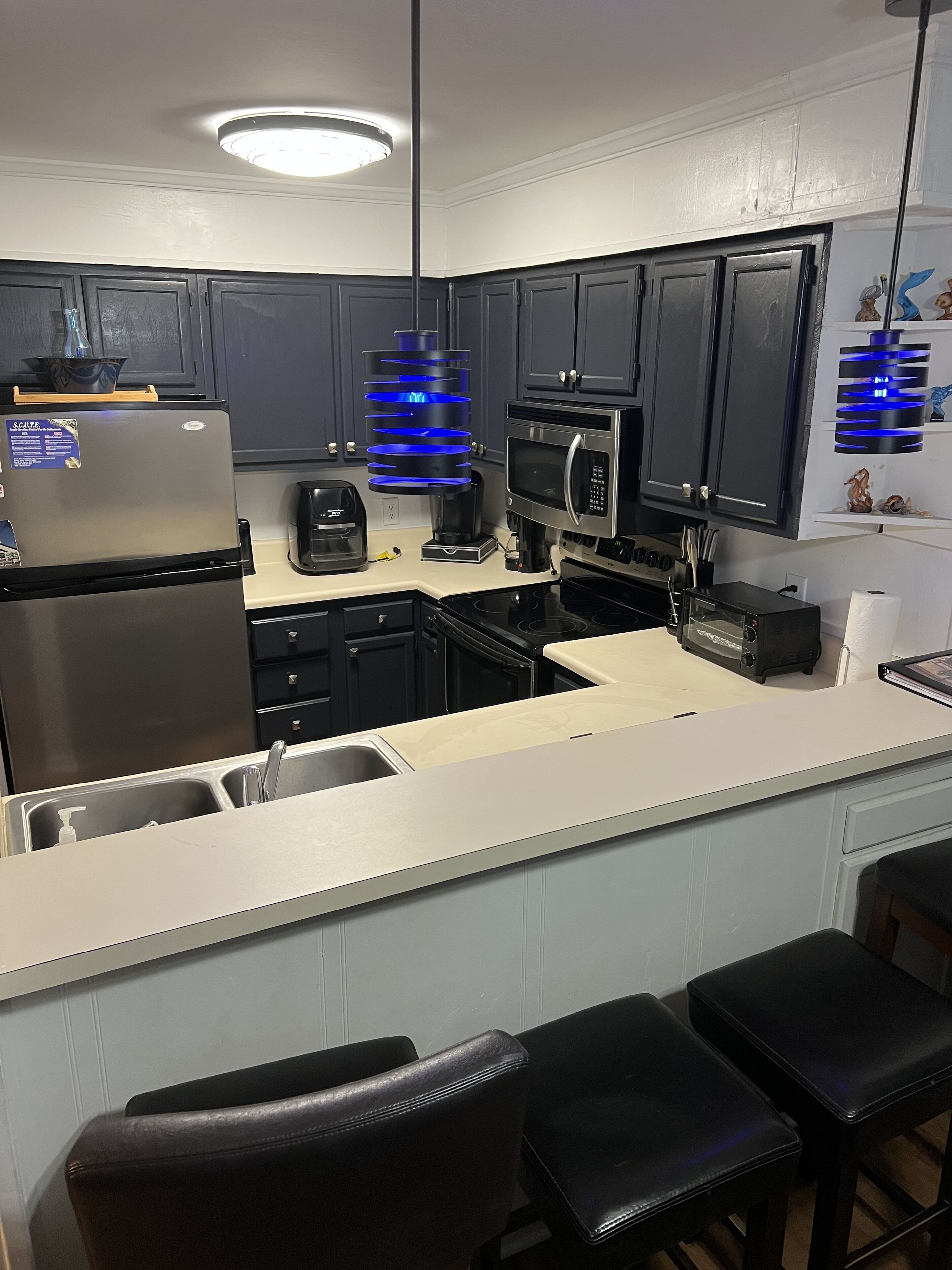A modern kitchen with a refrigerator, microwave, toaster, and a sink. There are black cabinets and a white countertop.