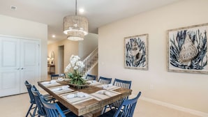 A dining area with a wooden table set for six, blue chairs, a hanging light fixture, and two framed artworks on the wall.