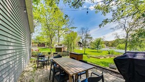 Eine Terrasse mit einem Holztisch und -stühlen, einem Grill und Blick auf eine grüne Rasenfläche und Bäume.