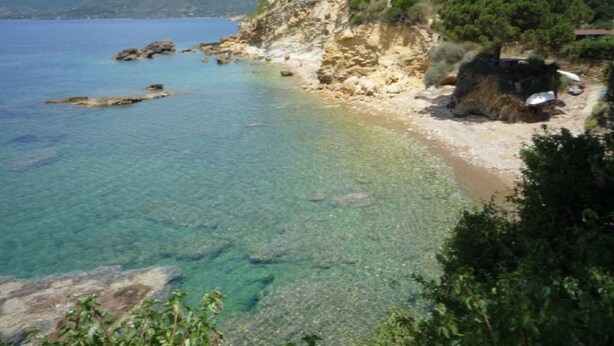 A coastal landscape with clear waters, rocky shorelines, and a hill with vegetation and buildings.