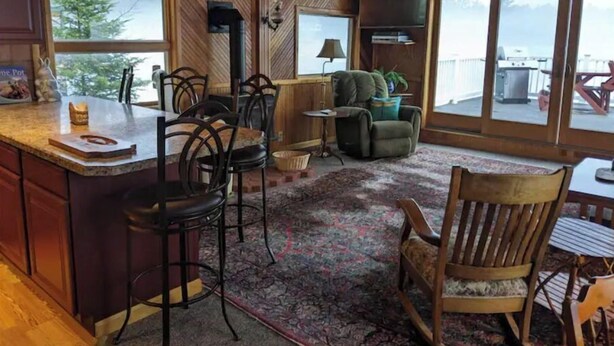 A cozy cabin interior with a wooden interior, a kitchen area with bar stools, a dining table, and a large window offering a view of the outdoors.