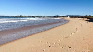 Praia extensa e arenosa com pegadas levando à água, céu azul límpido e linha costeira distante com edifícios.