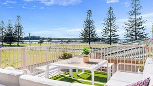 Varanda com vista para um parque e árvores, uma mesa branca e um sofá branco com almofadas.