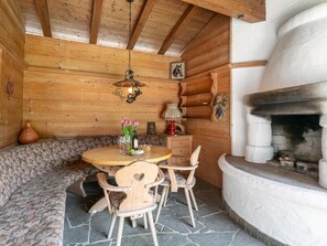 Un intérieur en bois avec un sol en pierre, une table ronde entourée de chaises, un banc, une cheminée et un luminaire suspendu.