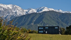 Un edificio moderno de color negro con ventanas amplias, rodeado por un campo y montañas al fondo.