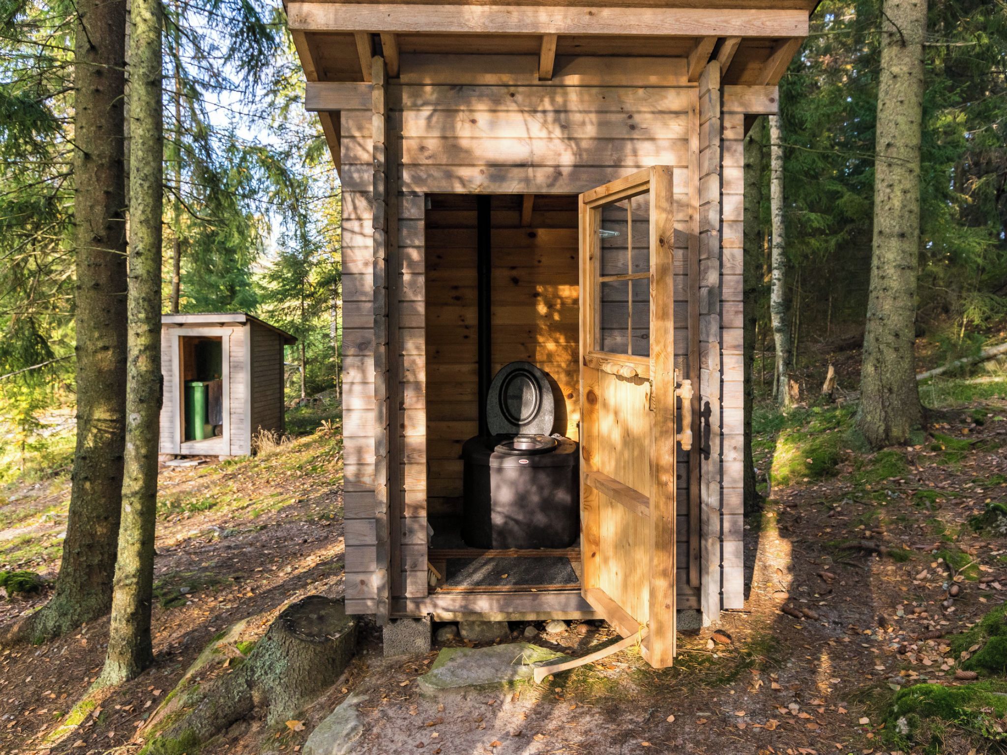 Una cabaña de madera con la puerta abierta, mostrando un inodoro y un cobertizo pequeño al fondo.