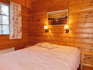Ein Zimmer in einer Holzhütte mit einem Bett, zwei Wandlampen, einem Fenster mit floralen Vorhängen und einem Bild an der Wand.