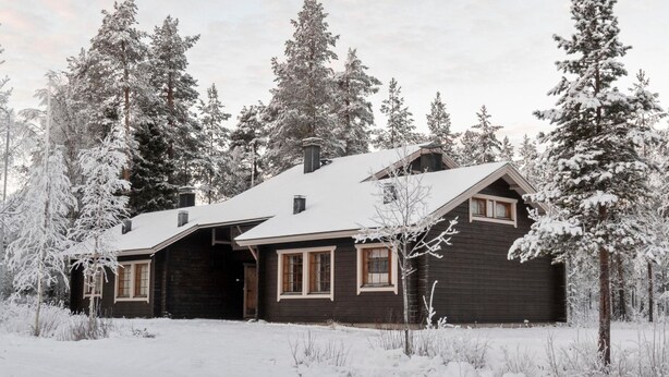 A cabin with a dark exterior, multiple windows, and a snow-covered roof, surrounded by tall, snow-laden trees. - Susitupa 6 by Interhome (Salla)