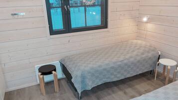 Ein kleiner, gemütlicher Raum mit einem Einzelbett, einem Hocker und einem Fenster mit Jalousien.