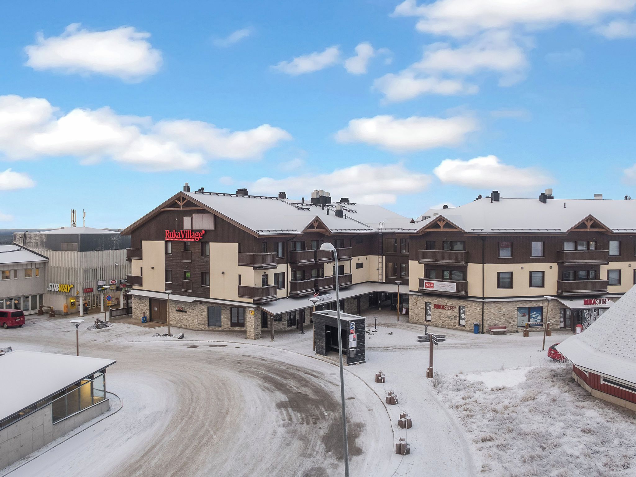 Un immeuble à plusieurs étages, avec un télésiège en arrière-plan, une entrée de restaurant Subway et un panneau indiquant le village de Ruka.