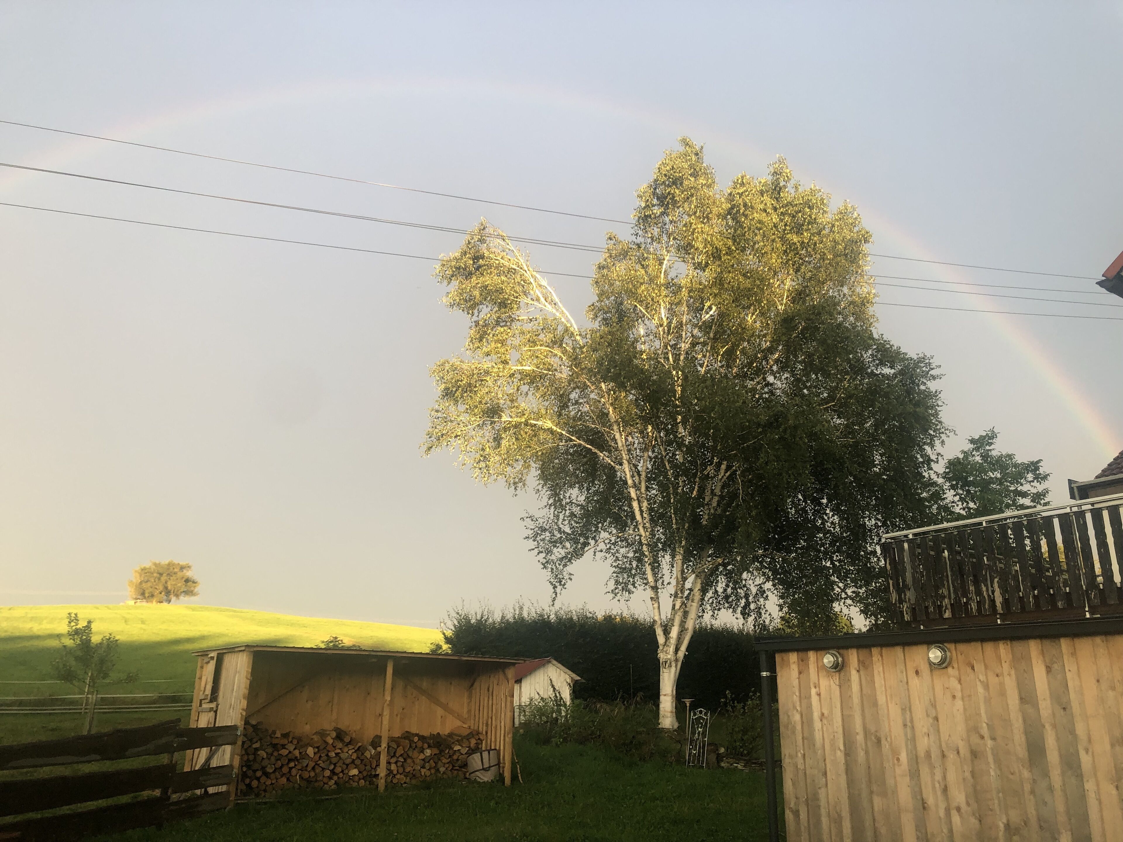 Un paisaje rural con un cobertizo de madera, un árbol y un arcoíris en el cielo.