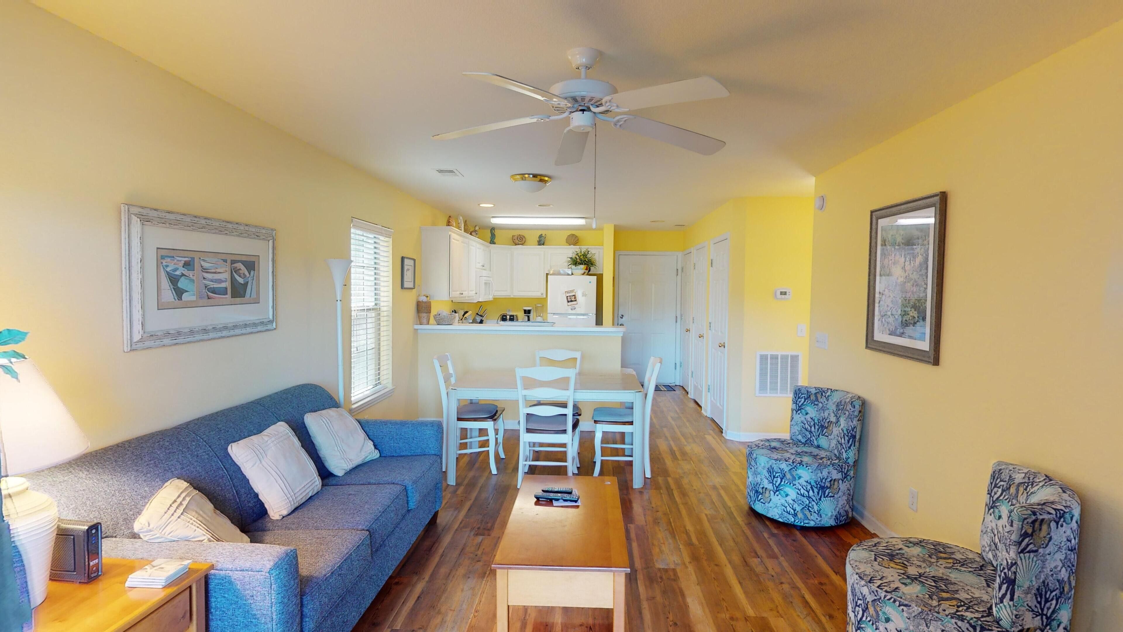 A compact living space with a sofa, a dining table with chairs, a ceiling fan, and a kitchen area with cabinets and a refrigerator.
