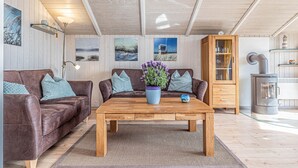 Un salón acogedor con un sofá de cuero marrón, una mesa de centro de madera y un jarrón azul con flores moradas. Se observan cuadros enmarcados en la pared, un aparador de madera y una estufa de leña.