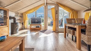 Un espacio habitable acogedor y de planta abierta con techo de madera, un televisor, una mesa de comedor con sillas de mimbre y un ventanal con cortinas amarillas.