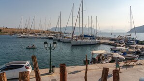 Un puerto deportivo con varios veleros amarrados, algunos coches estacionados cerca y una vista despejada del agua y montañas distantes.