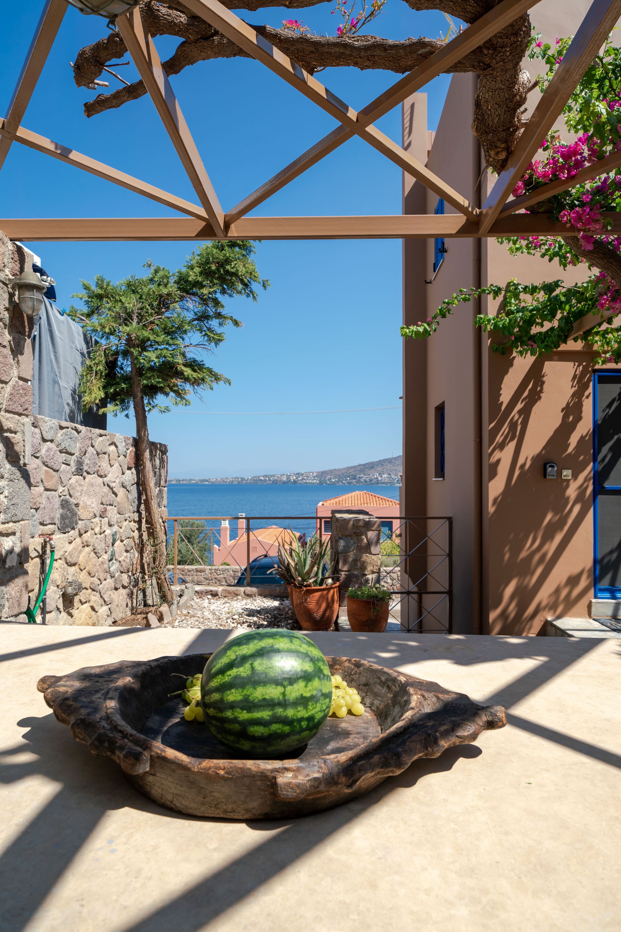 A stone wall, a tree, a wooden bowl with a watermelon, and a view of the sea.