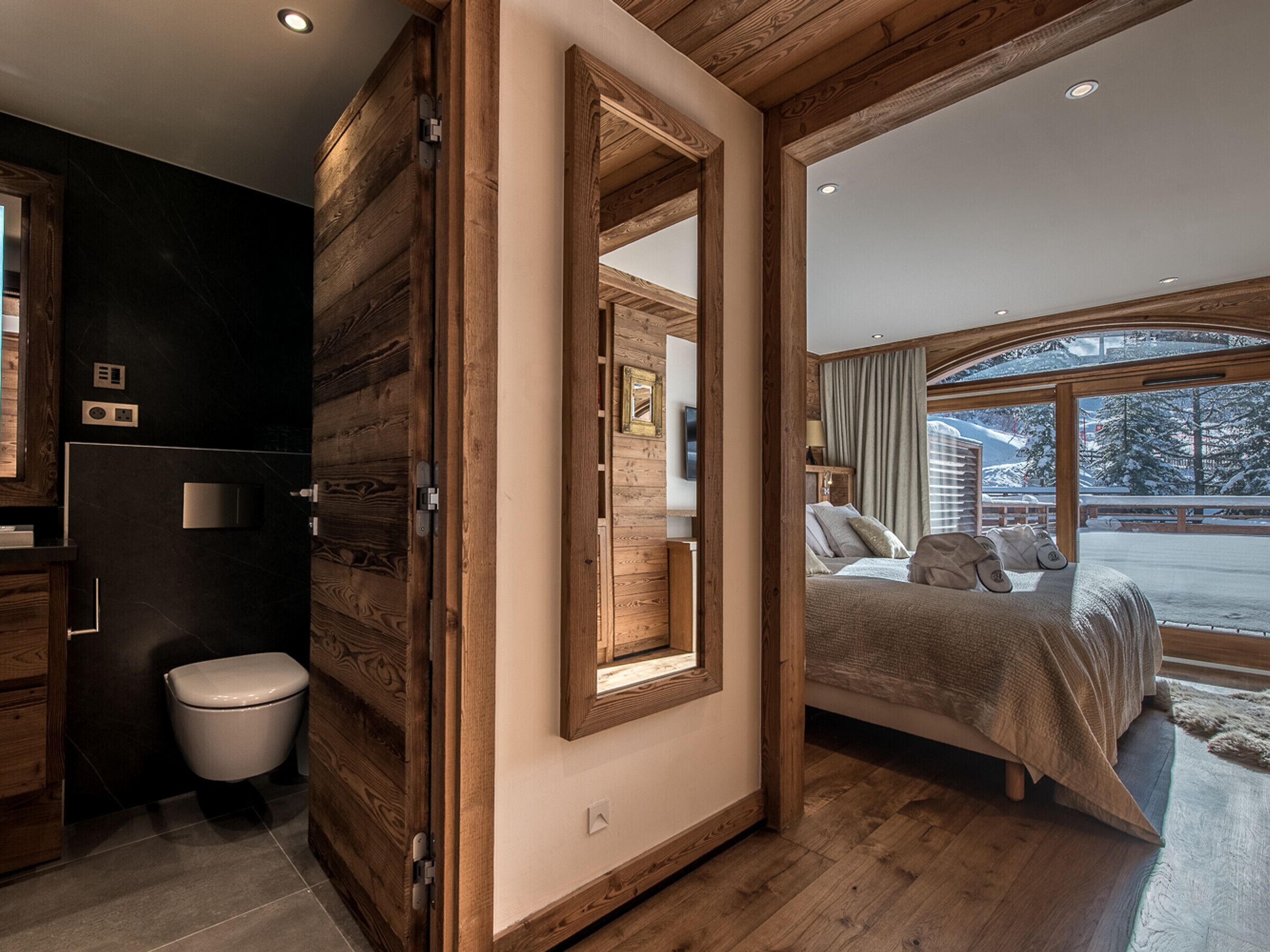 A modern bedroom with a wooden ceiling, a large bed, a view of a snowy landscape, and a bathroom with a wooden door.