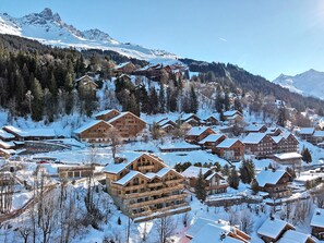 A snow-covered mountain resort with multiple wooden buildings, including a large lodge with balconies, set against a backdrop of snow-capped peaks and dense forest.