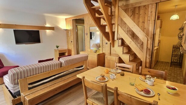 A cozy room with a wooden staircase, a sofa, a dining table with plates and cups, and a TV mounted on the wall.