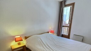 A small, neatly made bed in a room with white walls and a wooden floor. There are two bedside tables with lamps, one on each side of the bed. A window with curtains is visible in the background.