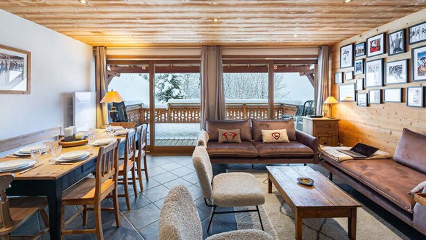 Une pièce chaleureuse avec un plafond en bois, un ensemble de canapés, une table à manger avec des chaises et une grande fenêtre donnant sur un paysage enneigé.
