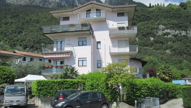Un edificio de varios pisos con balcones, coches aparcados y montañas al fondo.