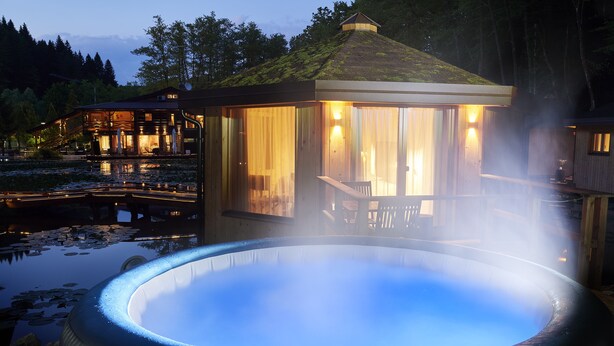A hot tub surrounded by a wooden gazebo with a view of a house and a pond.
