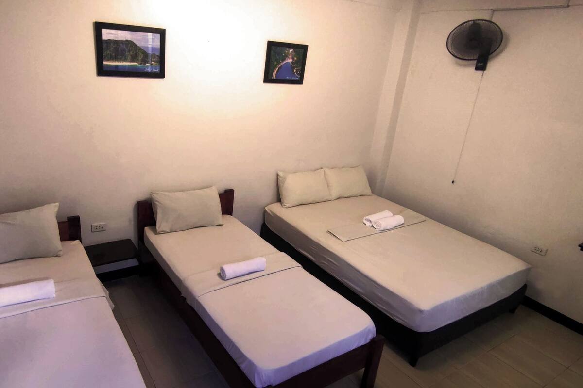 A small, compact hotel room with three single beds, each with a pillow and a rolled towel. There are two framed pictures on the wall, a small round mirror, and a shelf with a mirror.