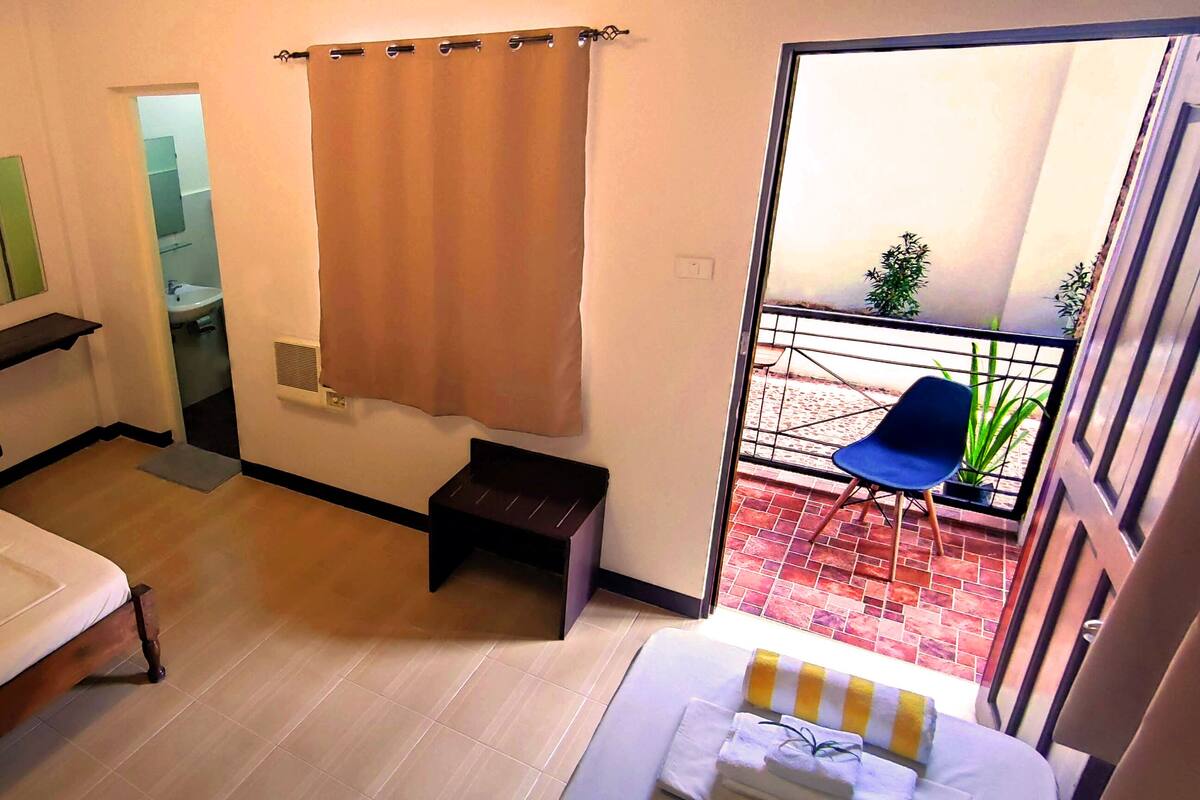 A hotel room with a bed, a small bench, a bathroom visible through an open door, and a balcony with a chair and plants.
