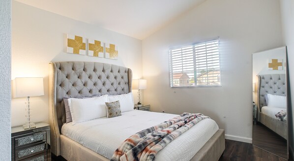 A bedroom with a tufted headboard, a large bed with white bedding, two bedside tables with lamps, a window with blinds, and a mirror on the wall.