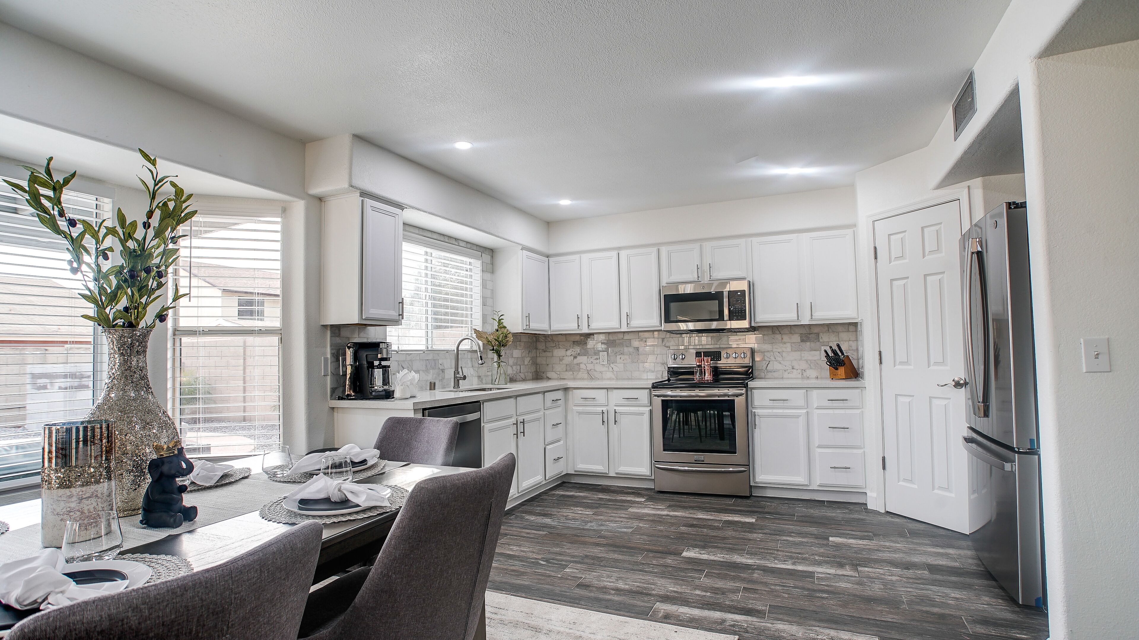 Una cocina moderna con armarios blancos, una zona de comedor con una mesa para cuatro personas y un amplio ventanal que proporciona luz natural.