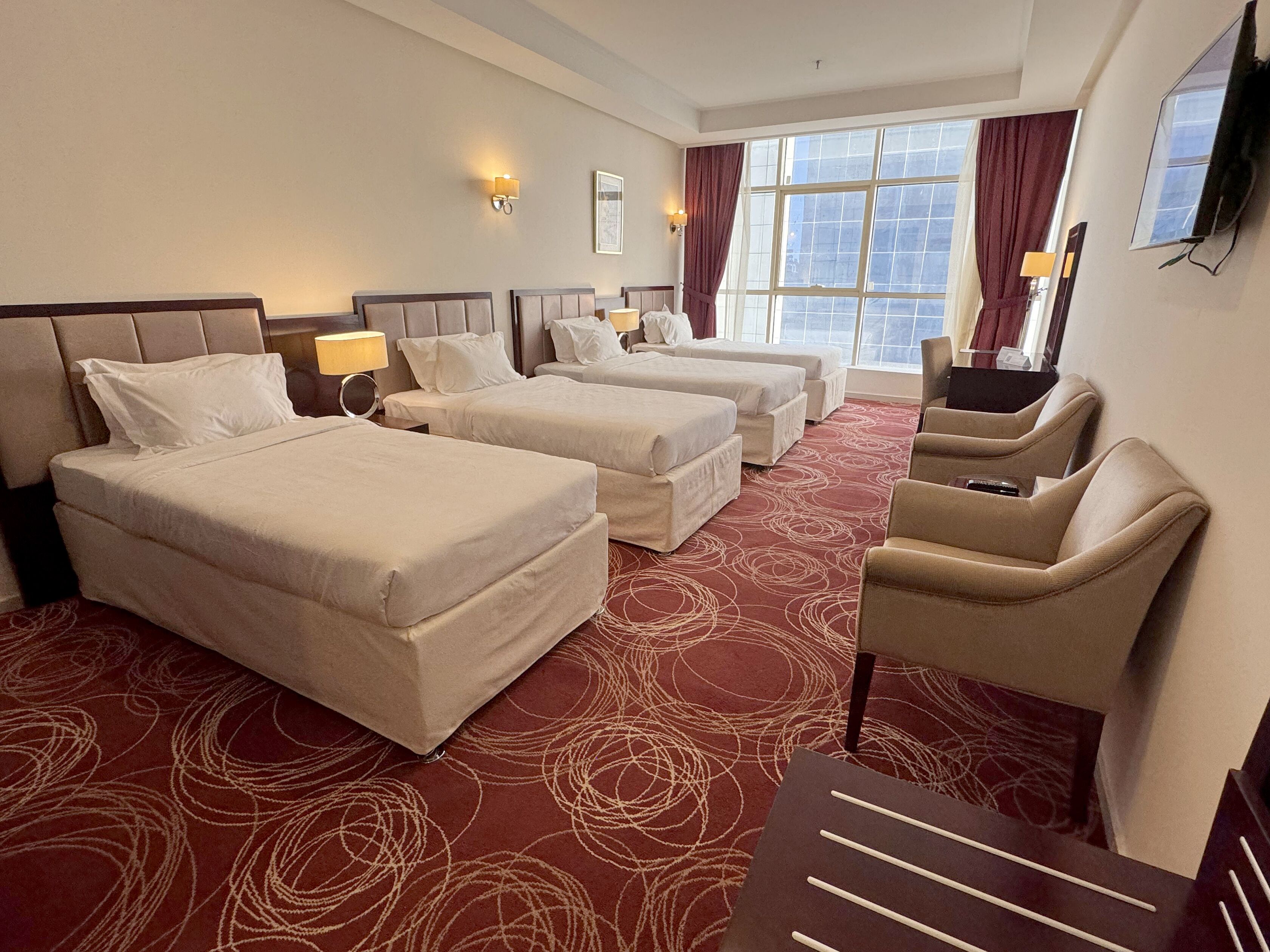 A hotel room with three beds, a TV, a desk, and a view of the city through large windows.