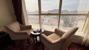 A hotel room with two beige armchairs, a small round table with a remote control, and a large window offering a city view. - Al Rayyan Makiya Towers Hotel (Makkah)