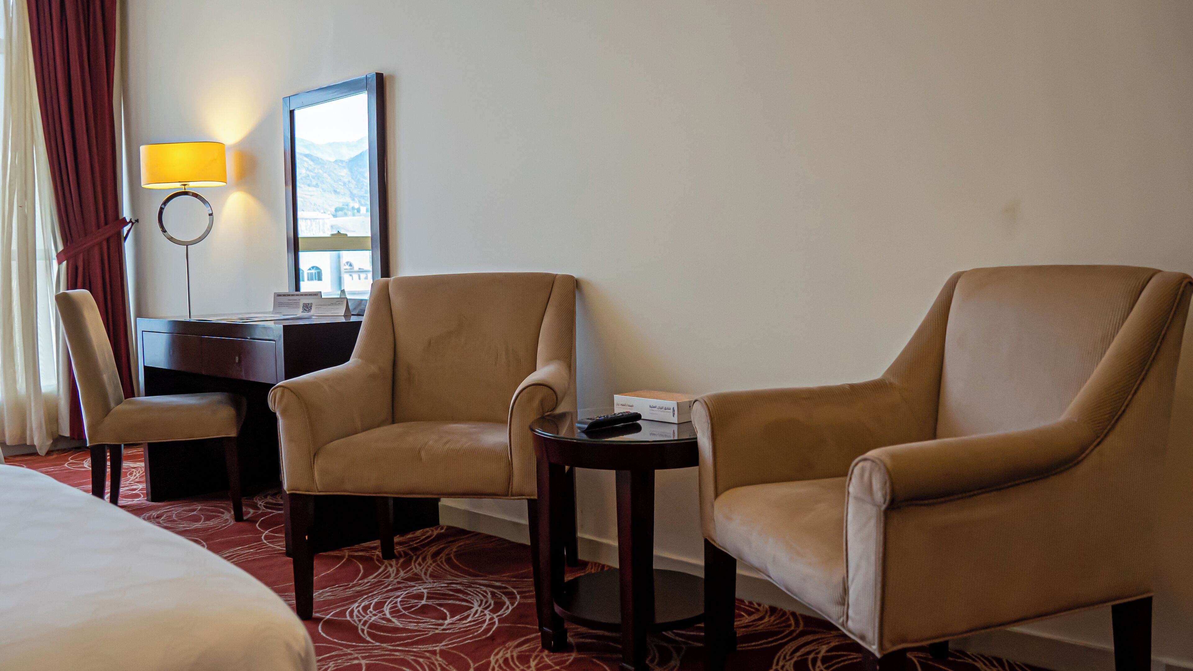 A hotel room with two armchairs, a desk, a lamp, and a bed.