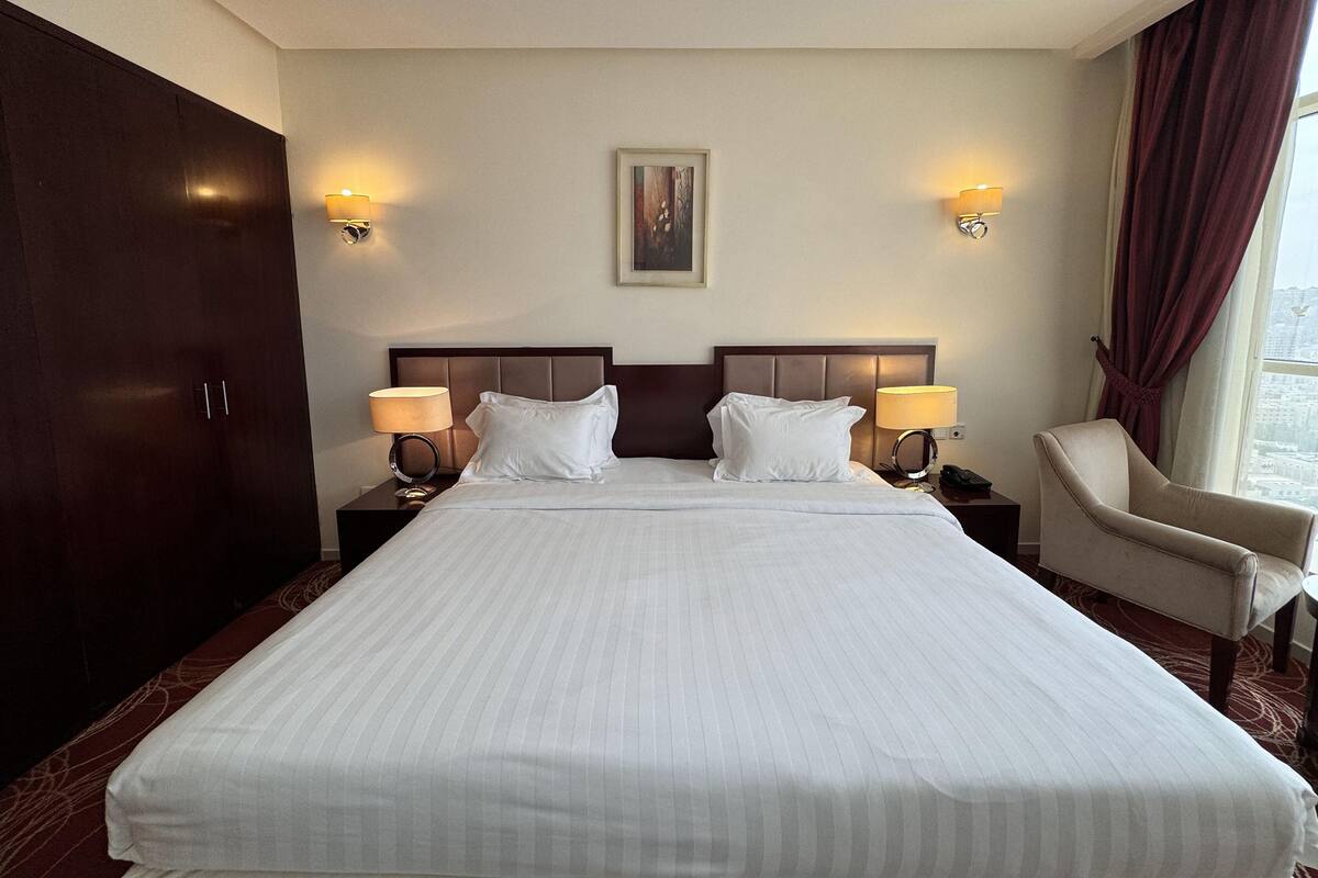 A hotel room with a large bed, two bedside lamps, a chair, a wardrobe, and a painting on the wall.