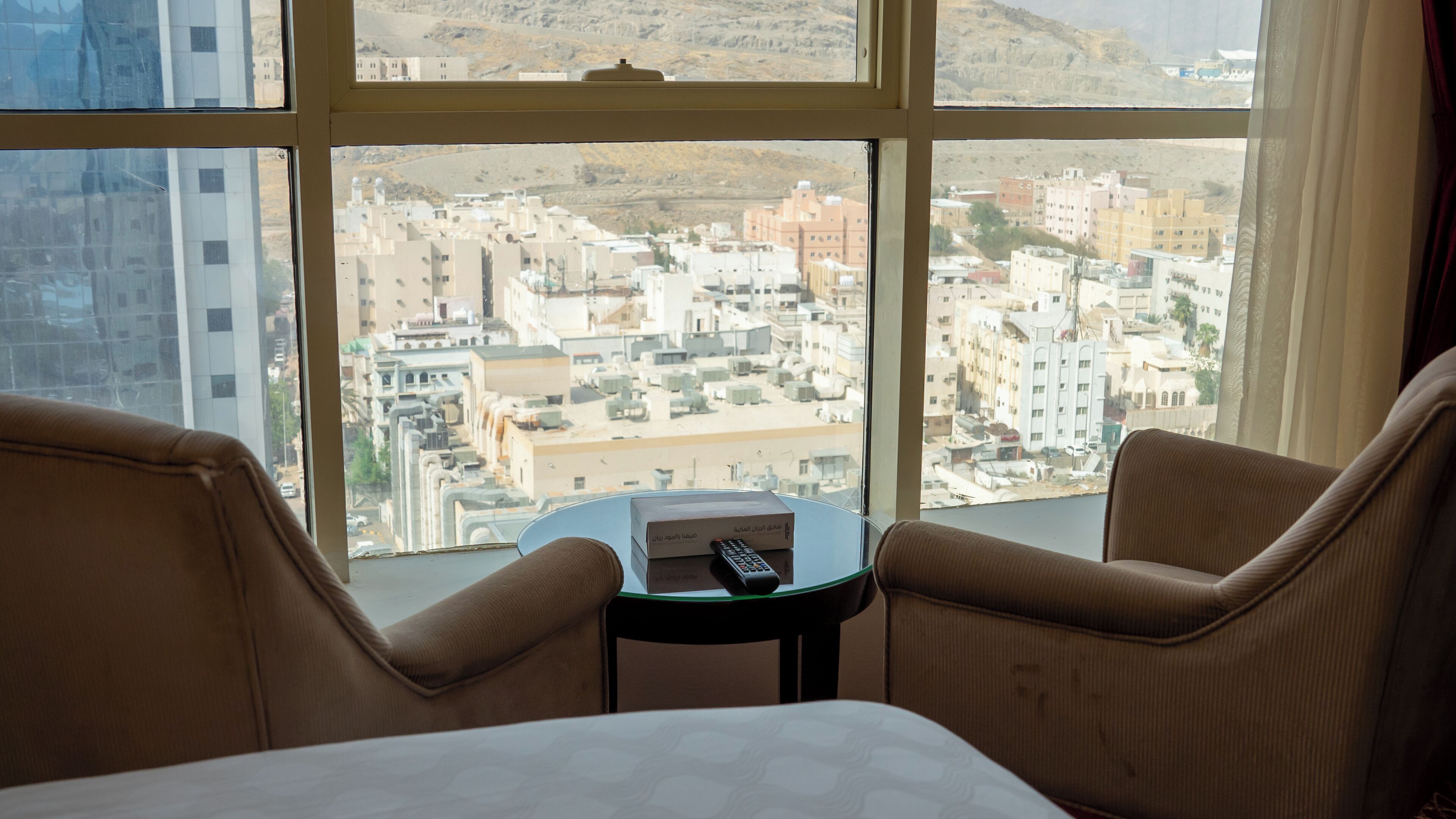 A hotel room with a large window offering a view of a cityscape. There are two brown armchairs and a small round table with a remote control on it.