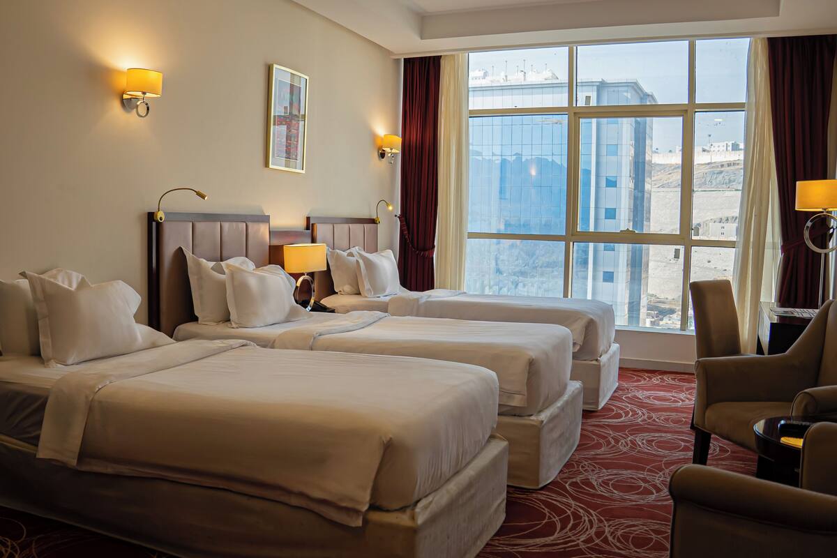 A hotel room with two beds, a desk, a chair, and a large window with a view of buildings.