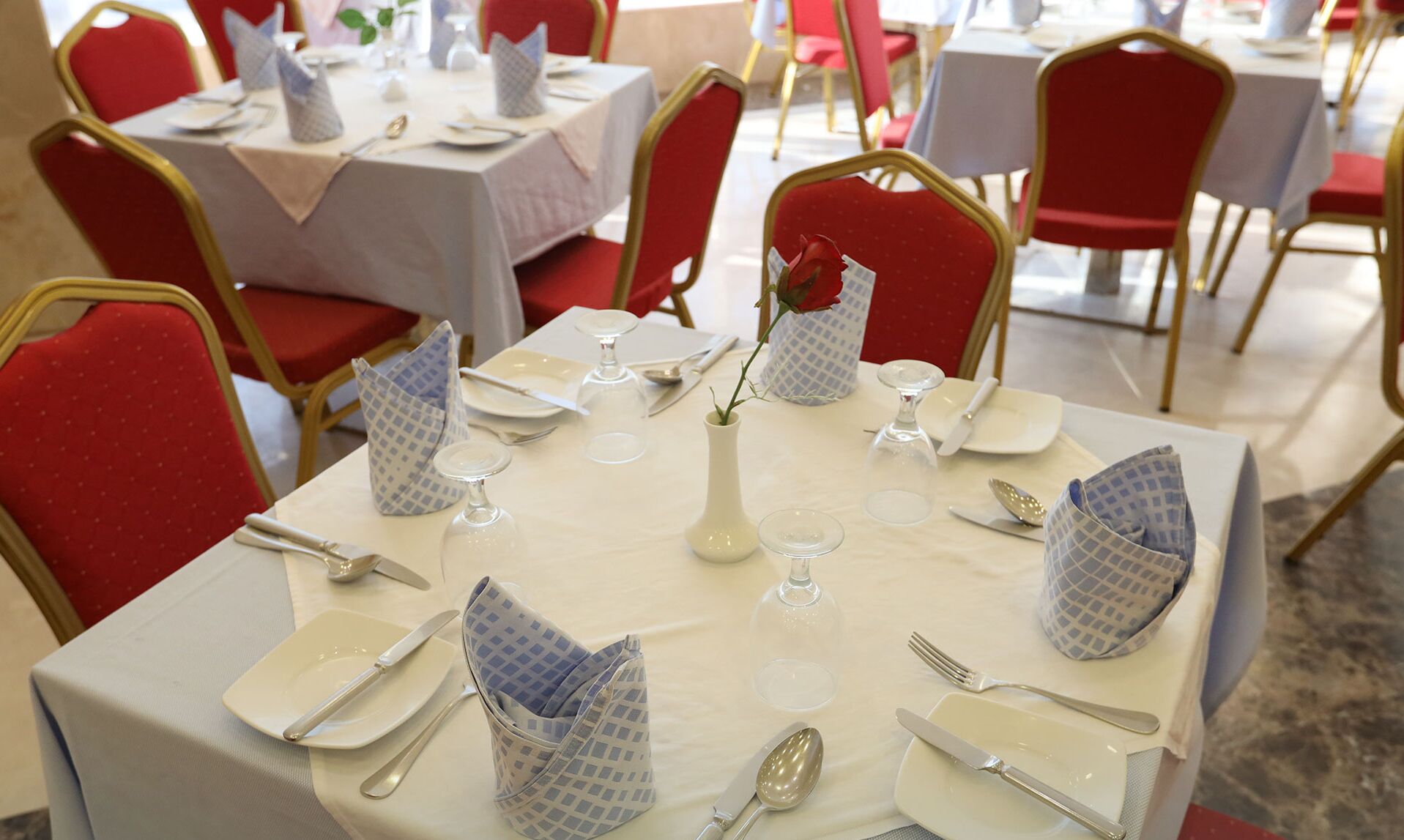 A dining table set with white tablecloths, red chairs, and blue napkin holders.