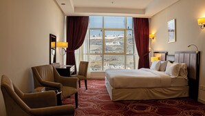 A hotel room with a large bed, two armchairs, a desk with a lamp, and a view of a cityscape through large windows. - Al Rayyan Makiya Towers Hotel (Makkah)