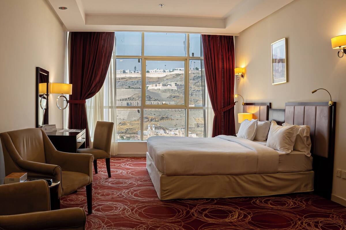 A hotel room with a large bed, two armchairs, a desk with a lamp, and a view of a cityscape through large windows.