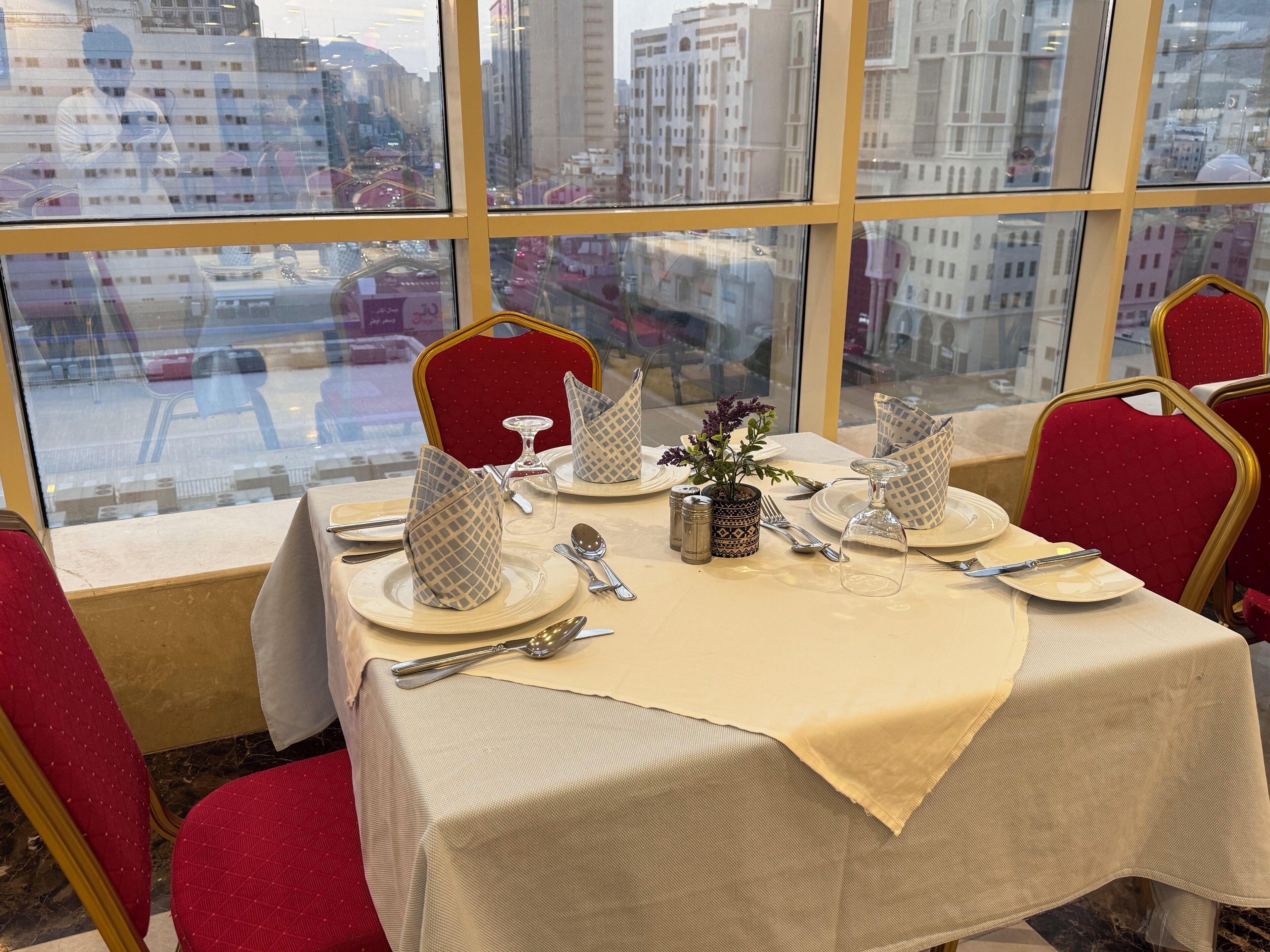 Una tavola imbandita con piatti, posate e un vaso di fiori, vista attraverso una finestra con uno skyline urbano sullo sfondo.