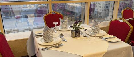 A dining table set with plates, cutlery, and a vase with flowers, viewed through a window with a cityscape in the background.