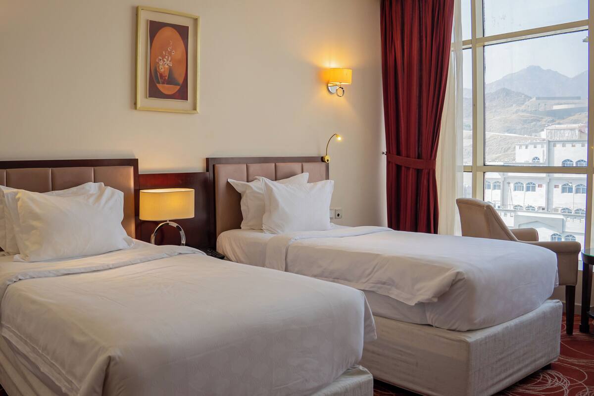 A hotel room with two beds, a painting on the wall, and a view of buildings through the window.