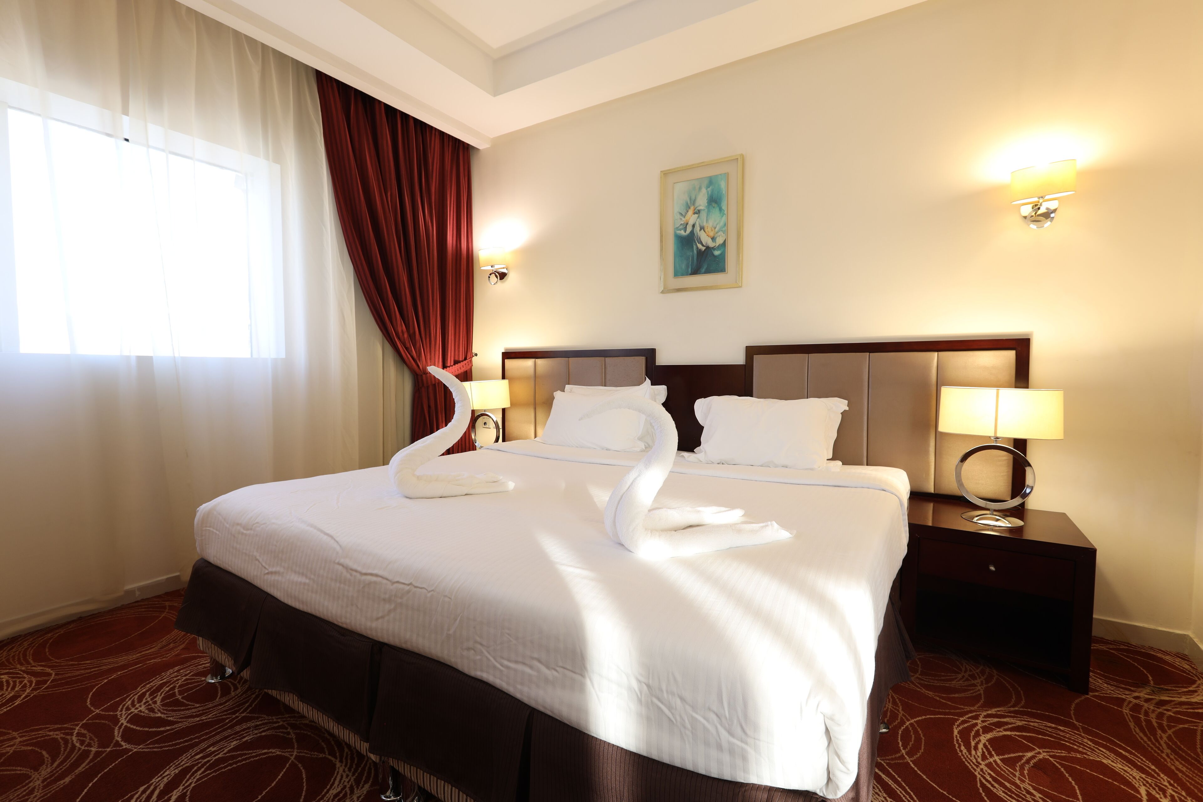 A hotel room with a large bed, two bedside tables with lamps, a painting on the wall, and a window with curtains.