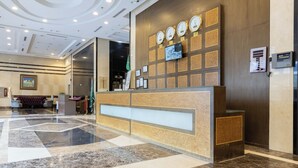 A hotel reception area with a marble floor, a reception desk, a wall-mounted clock, and a television screen. - Al Rayyan Makiya Towers Hotel (Makkah)