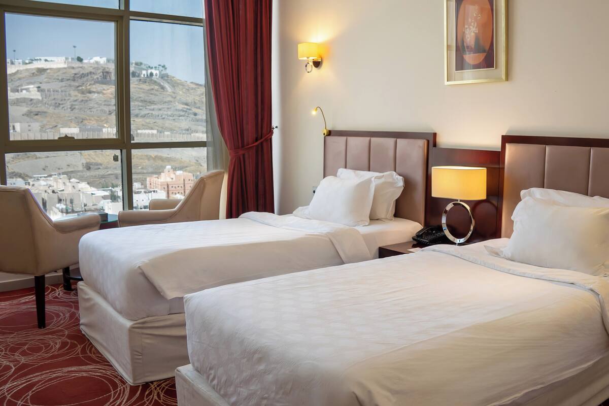 A hotel room with two beds, a desk, a chair, and a view of a cityscape.