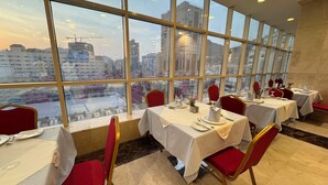 A restaurant with multiple tables and chairs, large windows offering a city view, and a clean, modern interior. - Al Rayyan Makiya Towers Hotel (Makkah)