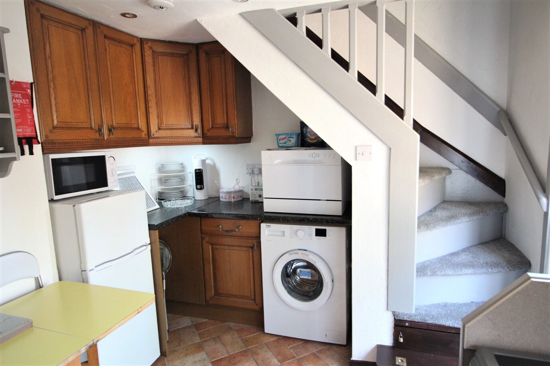 A compact kitchen with wooden cabinets, a microwave, a toaster, a washing machine, and a small table.