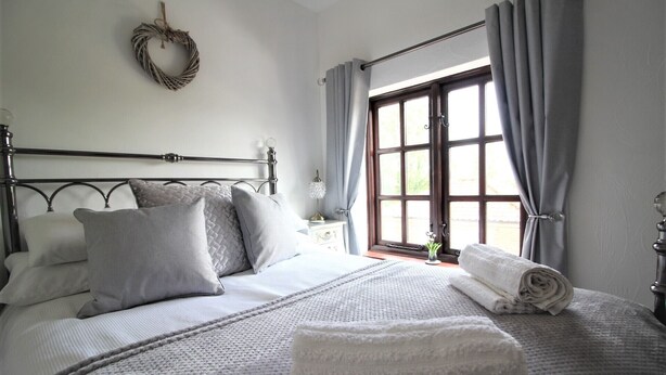 A neatly made bed with pillows and a blanket, a bedside table with a lamp, and a window with curtains. - Cosy Cottage with log burner, in the hart of the Norfolk countryside (Buxton)