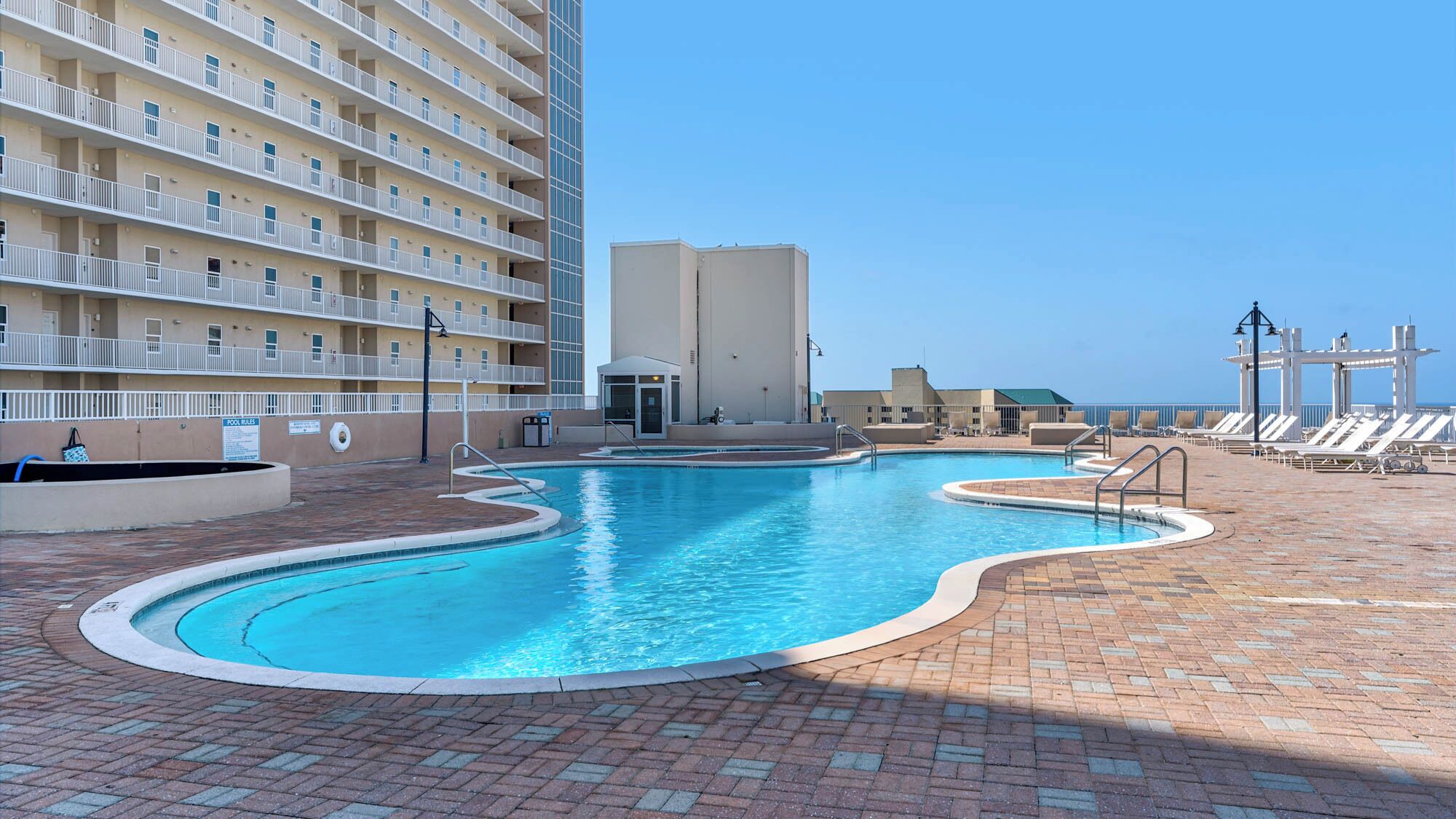 A swimming pool with a brick deck, a poolside lounge area, and a high-rise building in the background.